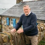 Pêcheur breton devant une petite maison en pierre avec volets bleus au bord de mer en Bretagne
